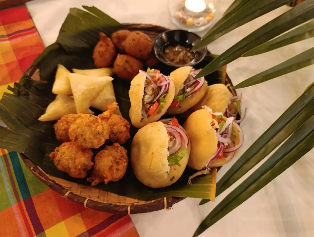 Assortiment apéritif créole avec petits pains garnis, acras, samoussas et sauces en panier décoré.