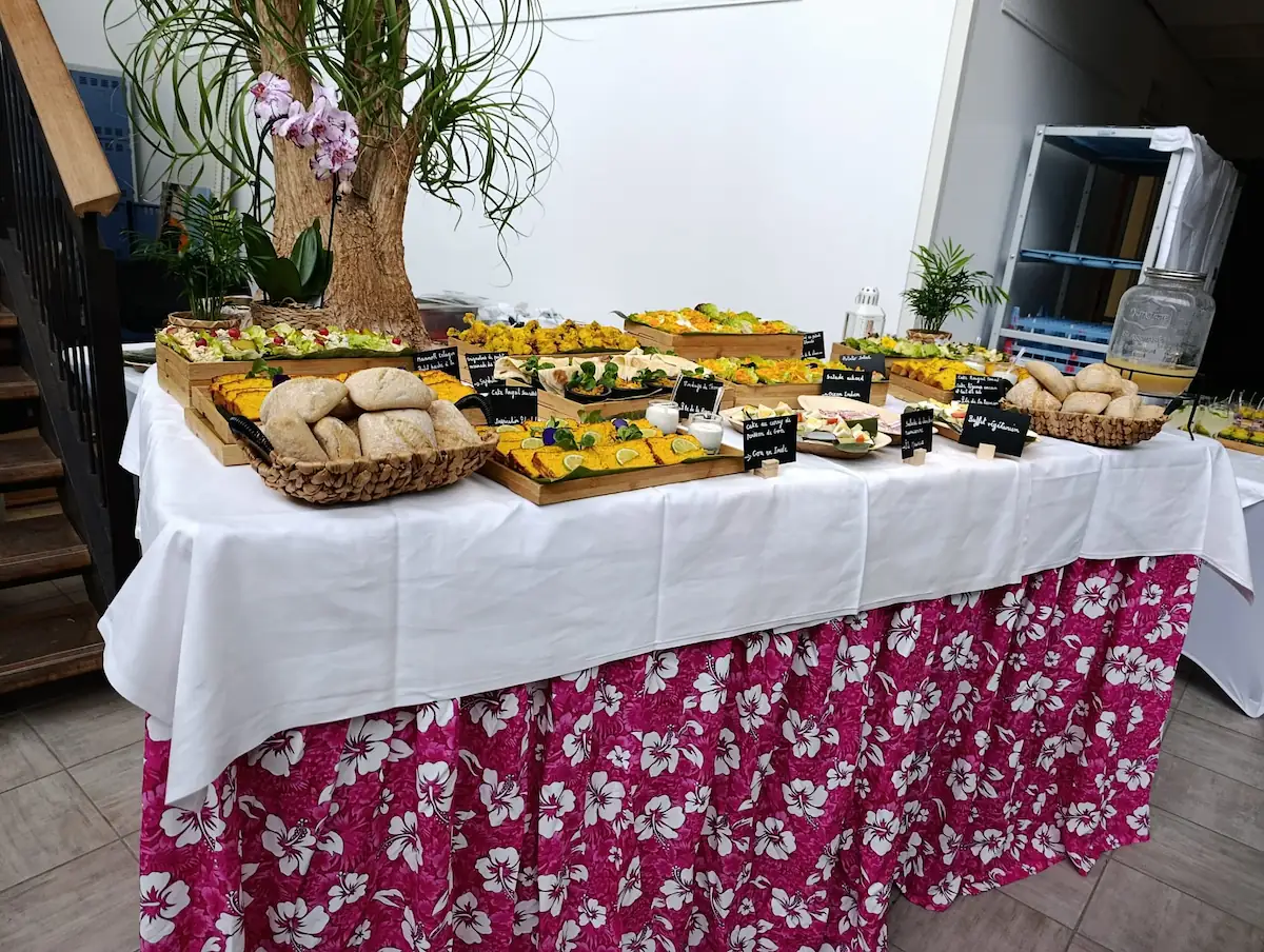 Buffet traiteur avec plateaux de mets variés, pains, plantes décoratives sur table avec nappe fleurie rose.