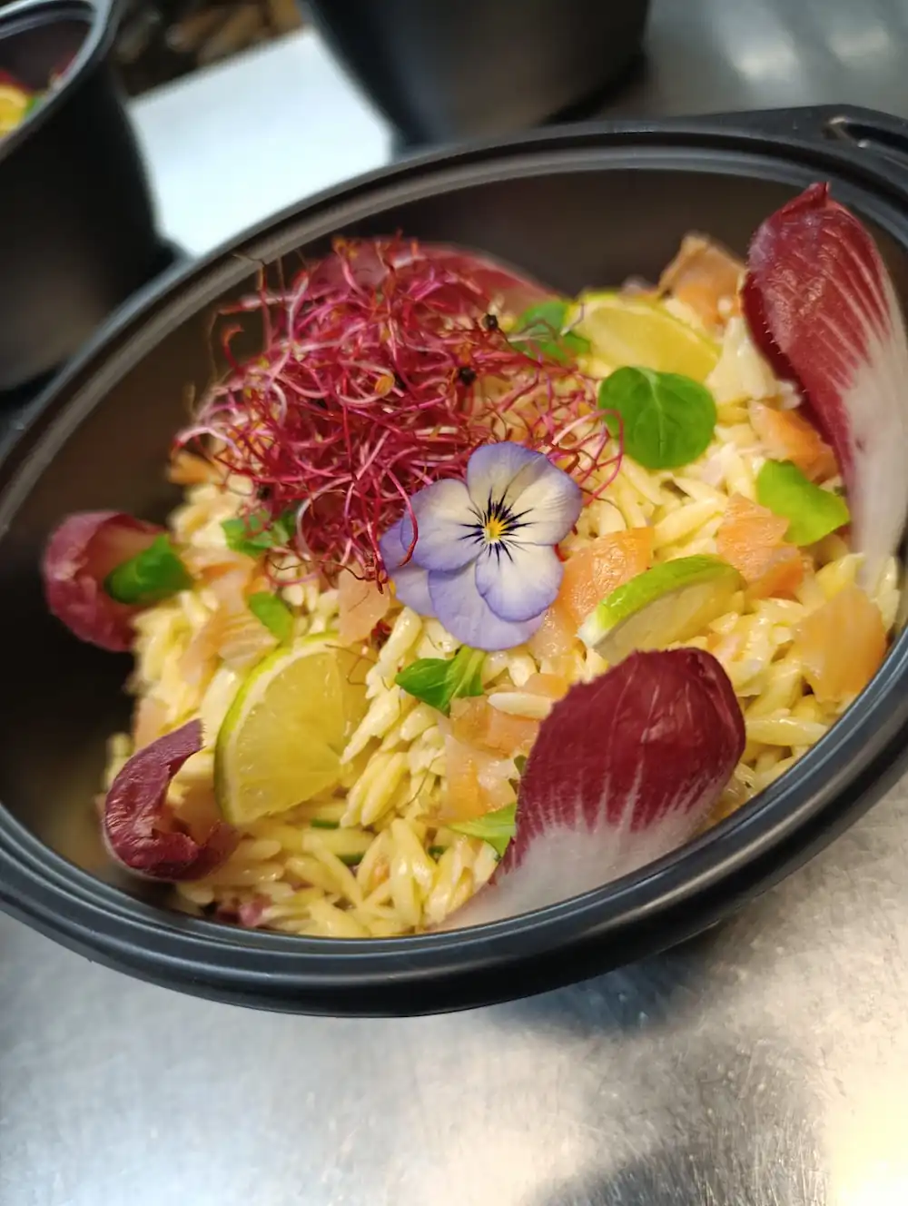 Salade avec pâtes orzo, rondelles de citron vert, feuilles rouges, fleurs comestibles et germes rougeâtres.